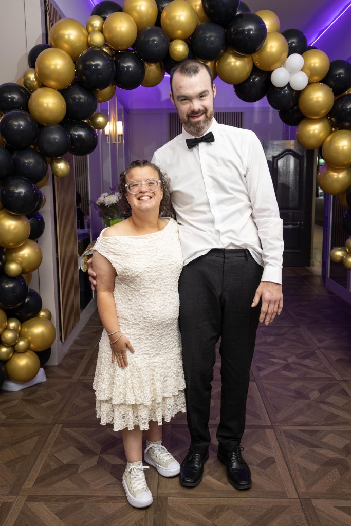 Two people stand in front of a balloon arch dressed for a formal occasion.
