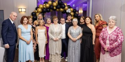 A group of thirteen people posing for a formal photograph in front of a black and gold balloon arch.