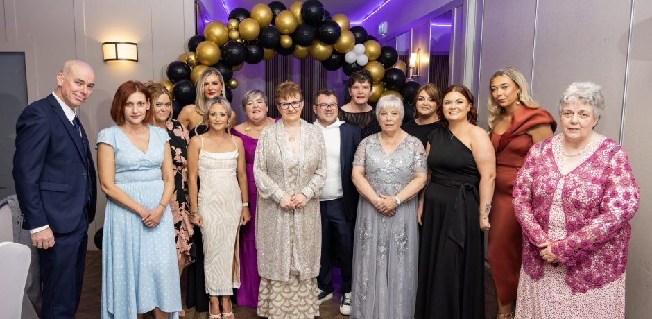 A group of thirteen people posing for a formal photograph in front of a black and gold balloon arch.