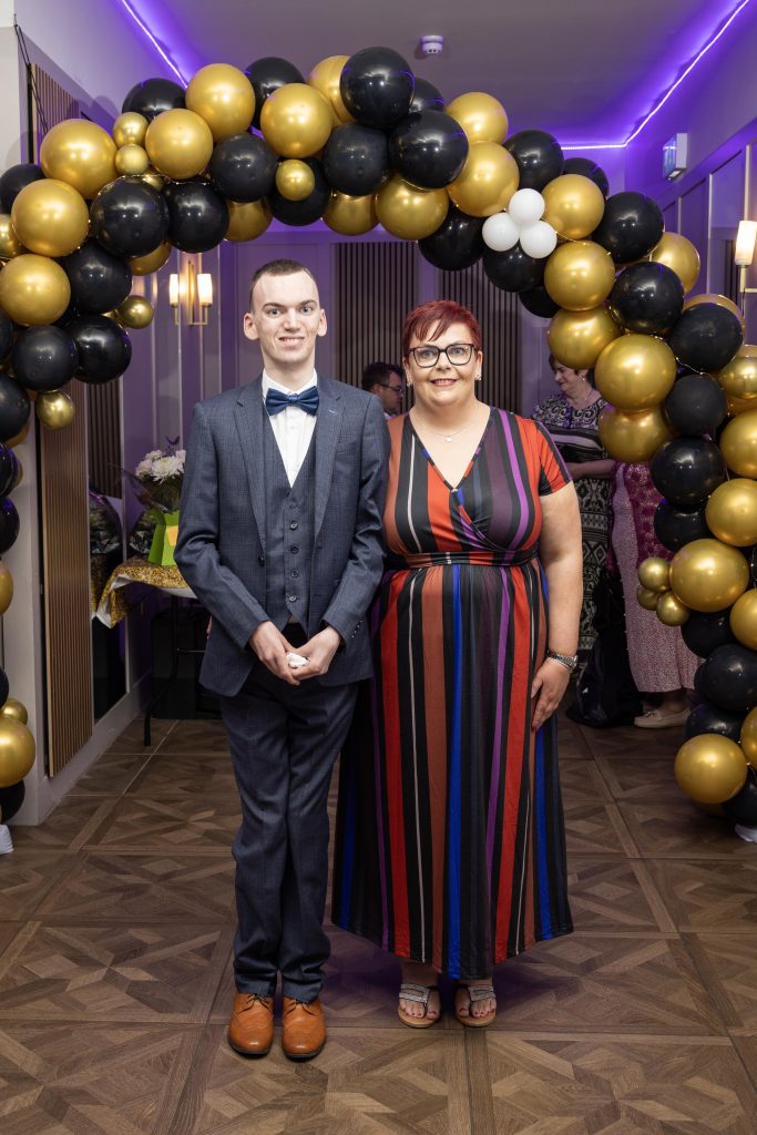 Two people stand in front of a balloon arch dressed for a formal occasion.