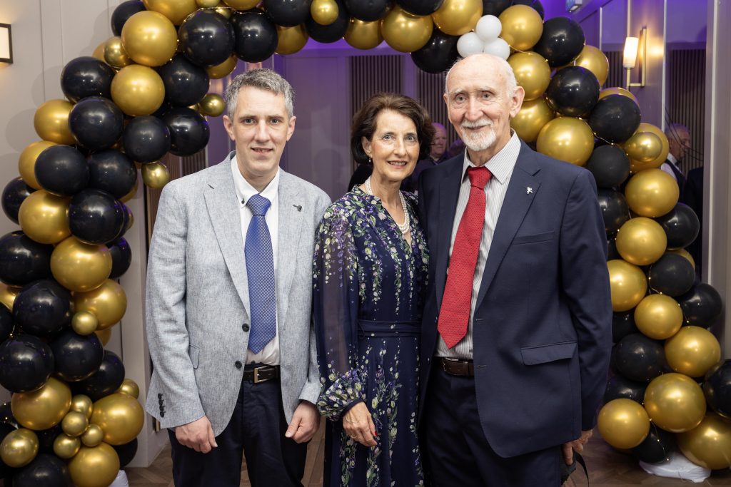 Three people stand in front of a balloon arch dressed for a formal occasion.