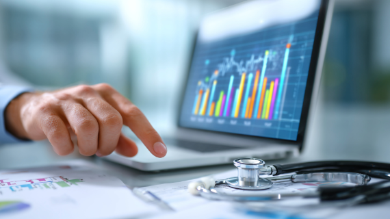 A person using a laptop show a dashboard and a stethoscope sitting beside it on the table