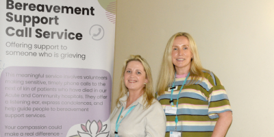 Two women standing in front of a Bereavement Support Call Service promotional banner at a table with purple folders.