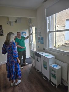 Two women stand in a room. They are looking at information sheets on the wall, and a number of different bins are lined up inside the room.