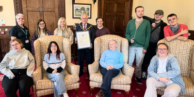A group of people including service users and staff members in a Mayor's Parlour setting. The Mayor is holding a framed certificate.