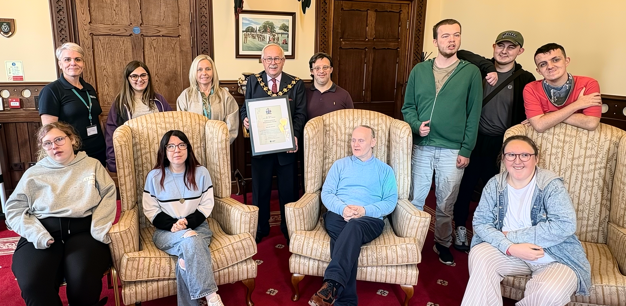 A group of people including service users and staff members in a Mayor's Parlour setting. The Mayor is holding a framed certificate.
