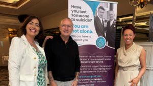 Three people standing in front of a banner which reads "Have you lost someone to suicide? You are not alone. We are here to support you."