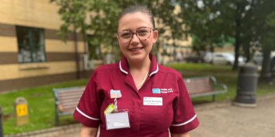 A smiling female healthcare worker in a maroon uniform standing outdoors in front of a building.