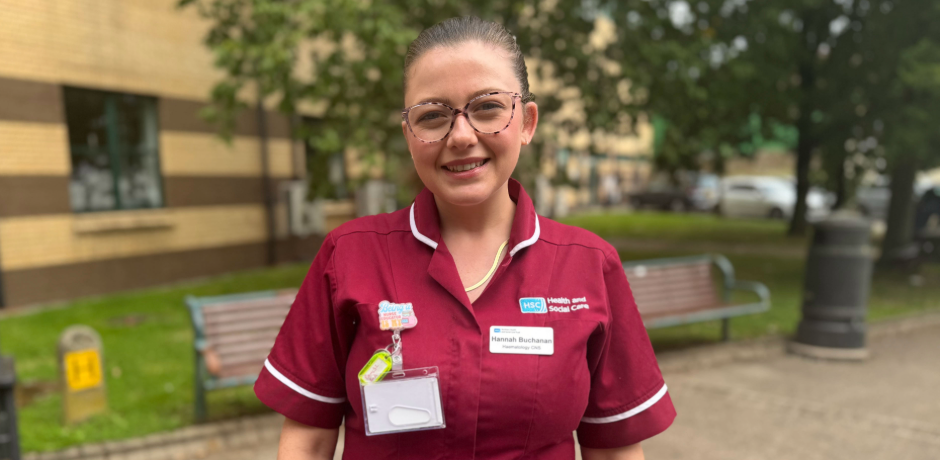 A smiling female healthcare worker in a maroon uniform standing outdoors in front of a building.