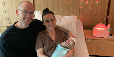 A mum and dad sit on a hospital ned together. Mum is holding a new born baby. A sensory type light is on the bedside table.