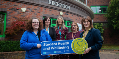 Five women stand with a red brick building behind them. They are holding a sign about student wellbeing.