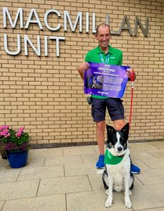 A man wearing a green Macmillan t-shirt stands outside the Macmillan Unit with his collie dog Charlie in front of him. Charlie is wearing a green Macmillan bandana. Ivan is holding a poster about the BBC Make a Difference awards.