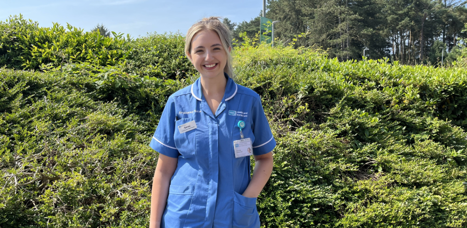 A nurse in a blue tunic stands outside on a sunny day with a green hedge behind.
