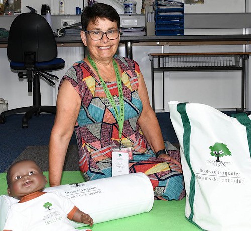 Belinda Simpson has been involved with Roots of Empathy at Ballycraigy Primary School in Antrim since 2013 and she’s now celebrating 10 years as an instructor. Belinda Simpson sitting on a mat inside a classroom with a dolls and a bag which has the Roots of Empathy logo on it.